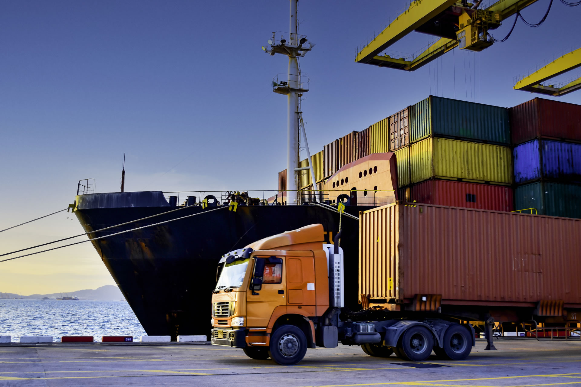 A sunny shipping terminal featuring large blue gantry cranes towering over high stacks of colorful intermodal shipping containers ready for export