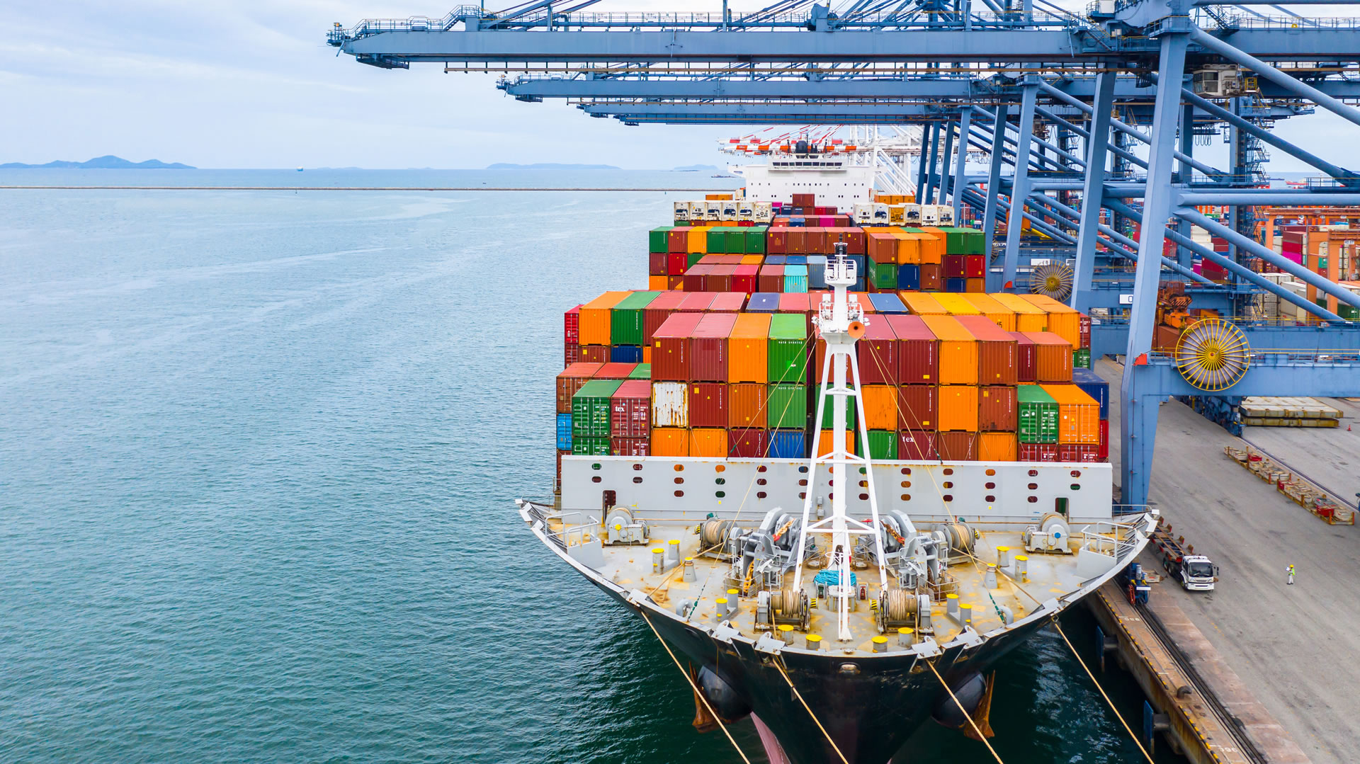An orange semi-truck and container chassis parked on the dock alongside a large cargo vessel, highlighting the transition between maritime and road logistics